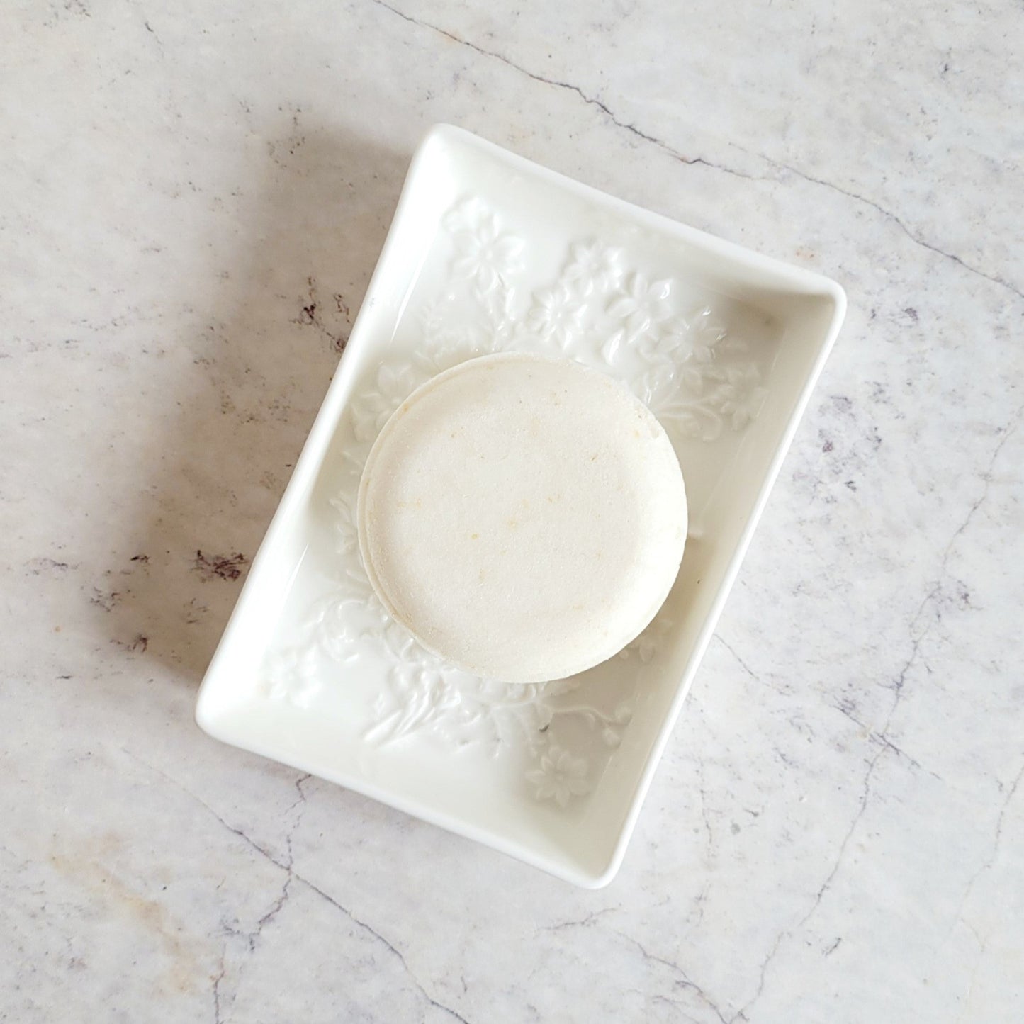 Solid Shampoo Bar, round, white disk with specs of ground oatmeal. Shampoo bar is in a white ceramic soap dish,