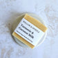 suare golden soap bar with label. sitting on a ceramic round soap dish